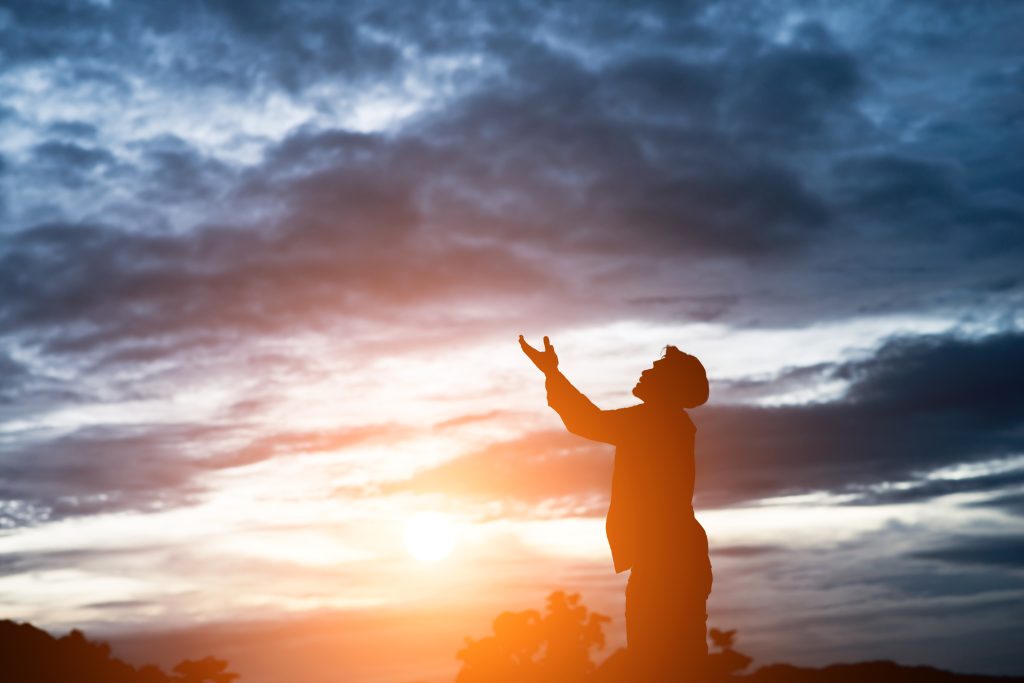 Silhueta de uma pessoa com os braços erguidos em direção ao céu durante o pôr do sol, transmitindo uma sensação de fé, esperança e conexão espiritual, com nuvens dramáticas no fundo e tons dourados iluminando a cena.