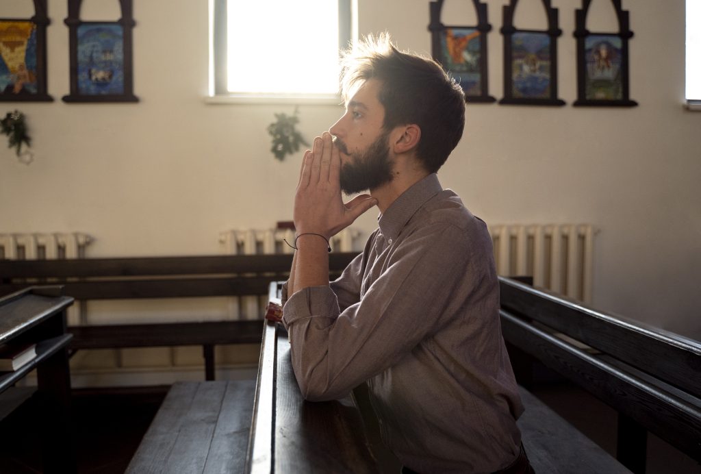 Homem sentado em um banco de igreja, com as mãos unidas em posição de oração e expressão reflexiva, iluminado pela luz do sol que entra pela janela, transmitindo um sentimento de fé, introspecção e busca por paz interior.