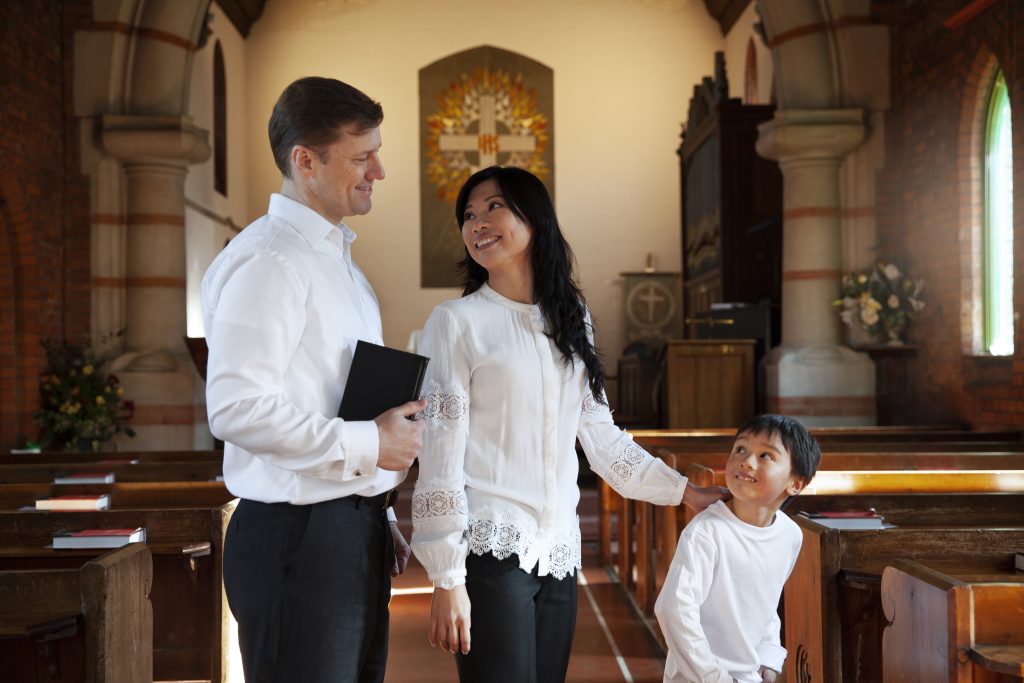 Texto alternativo: Família sorridente dentro de uma igreja, vestindo roupas brancas, com o pai segurando uma Bíblia, a mãe tocando carinhosamente no filho e o ambiente transmitindo fé, harmonia e conexão espiritual.