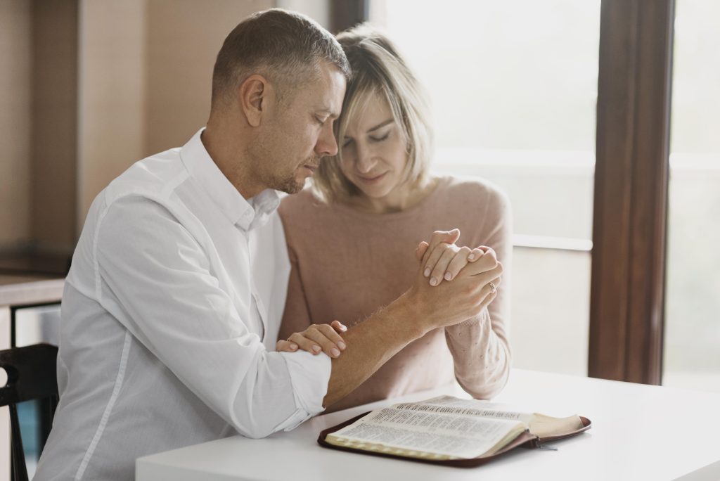 Casal sentado à mesa, de mãos dadas e olhos fechados em oração, com uma Bíblia aberta à frente, transmitindo fé, união e busca por restauração no relacionamento sob uma luz suave e acolhedora.