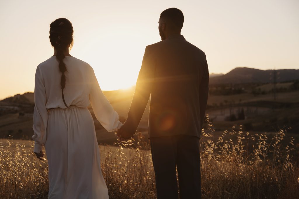 Casal de mãos dadas observando o pôr do sol em um campo aberto, com roupas elegantes e um ambiente dourado ao redor, simbolizando amor, união e conexão espiritual em um cenário tranquilo e inspirador.